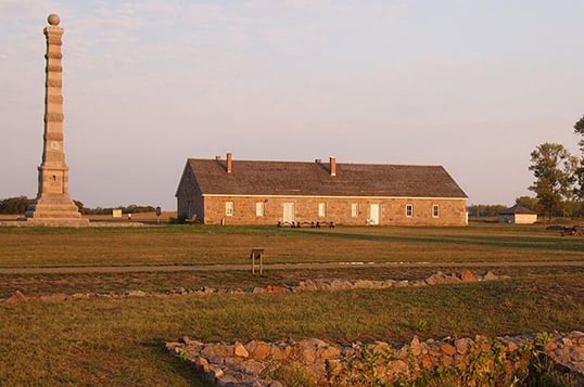 View of Fort Ridgey with stone foundation ruins in the foreground, a one-story building with two doors and six windows in the middleground next to a stone pillar, and a grey sky in the background.