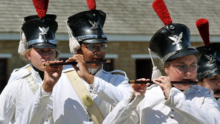 Photo of historic re-enactors in a fife and drum corps wearing black hats with red feathers and metal emblems