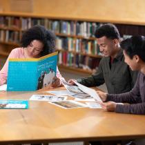 Three teenagers sit at a table looking at papers and writing notes.